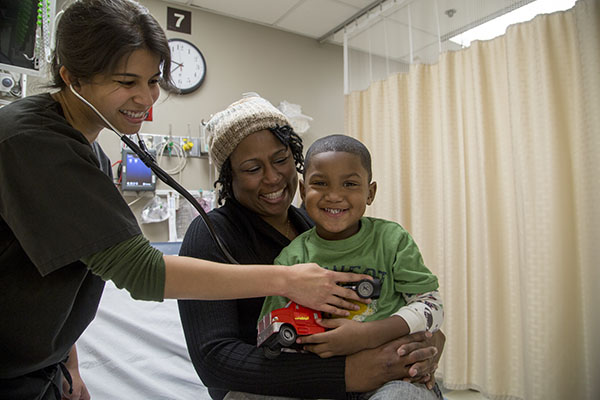 A woman holding a stethescope to a young, Black male holding a toy truck while he sits in his mother's lap.