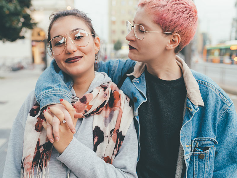 A non-binary person with short pink hair, glasses and dark lipstick with their arm around a female-presenting person with their hair pull back and glasses.