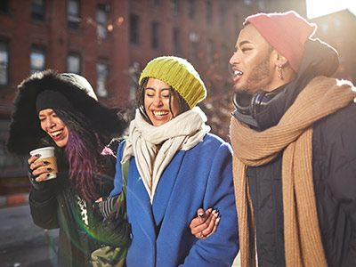 Two dark-skinned female-presenting people with male-presenting person wearing eyeliner and stubble, all wearing winter coats and hats walking and laughing 