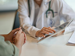 A doctor gesturing to a tablet device across the table from a patient.